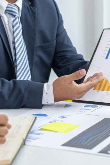 Two people in business attire discuss graphs and charts at a table, with one person holding a clipboard displaying bar and line graphs to better understand customer behaviour in their business.