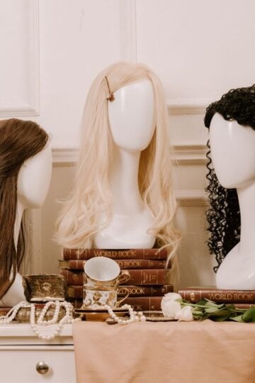 Three mannequin heads with wigs—brown, blonde, and black curly—are displayed among stacked “World Book” volumes, pearls, a teacup, and a white rose on a beige tablecloth—a charming tableau perfect for anyone seeking a beginner’s guide to glueless wigs or exploring different wig styles.