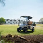 A silver golf buggy is parked on a grassy area with rocks in the foreground and white residential buildings in the background, making it an ideal setting for family golf or those exploring starter golf equipment.