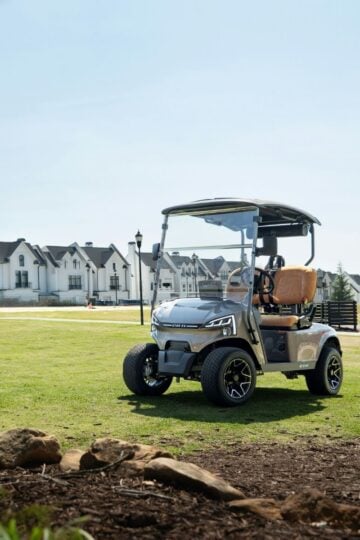 A silver golf buggy is parked on a grassy area with rocks in the foreground and white residential buildings in the background, making it an ideal setting for family golf or those exploring starter golf equipment.