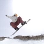 Person in goggles and winter gear, sporting mum-tested kids' snowboard boots that guarantee a perfect snowboard boots fit, snowboarding mid-air above a snowy slope with trees in the background under a bright sky.