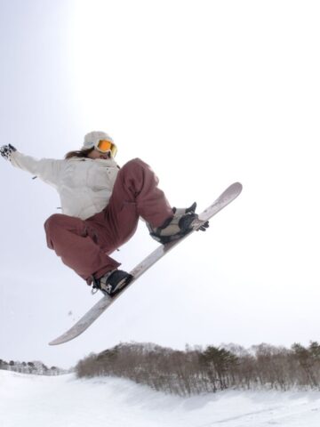 Person in goggles and winter gear, sporting mum-tested kids' snowboard boots that guarantee a perfect snowboard boots fit, snowboarding mid-air above a snowy slope with trees in the background under a bright sky.