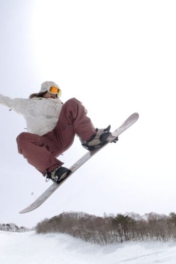 Person in goggles and winter gear, sporting mum-tested kids’ snowboard boots that guarantee a perfect snowboard boots fit, snowboarding mid-air above a snowy slope with trees in the background under a bright sky.
