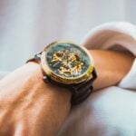 A close-up of a wrist wearing a mechanical watch with visible gears, a gold bezel, and a brown leather strap, showcasing the timeless style that defines luxury watches and their generational appeal.