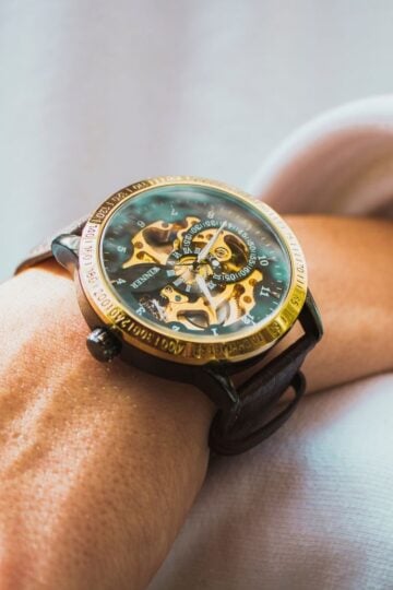 A close-up of a wrist wearing a mechanical watch with visible gears, a gold bezel, and a brown leather strap, showcasing the timeless style that defines luxury watches and their generational appeal.