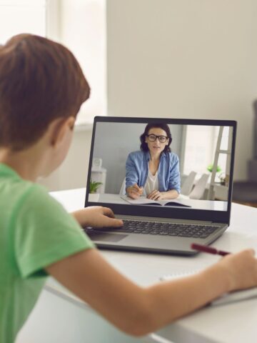 A boy sits at a desk watching a woman on a laptop screen during an online lesson, with a notebook and pen in front of him, as he explores practical strategies for effective home schooling.