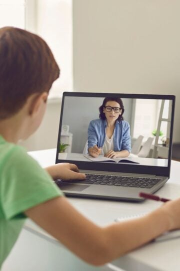 A boy sits at a desk watching a woman on a laptop screen during an online lesson, with a notebook and pen in front of him, as he explores practical strategies for effective home schooling.