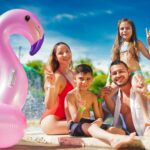 A family of four poses by a swimming pool with a large pink inflatable flamingo, all smiling and making peace signs on a sunny day.
