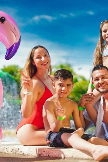A family of four poses by a swimming pool with a large pink inflatable flamingo, all smiling and making peace signs on a sunny day.