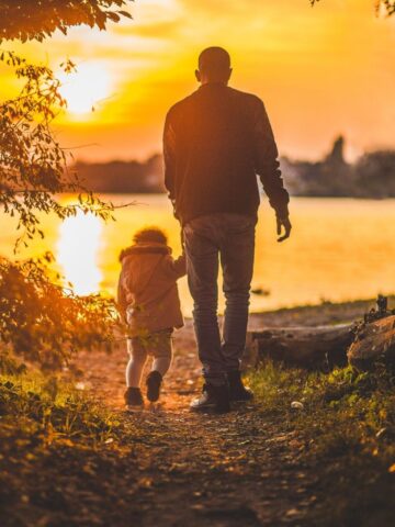 An adult and a child walk hand in hand towards a lake at sunset, surrounded by trees and warm orange light-capturing a perfect moment for those seeking family photography tips.