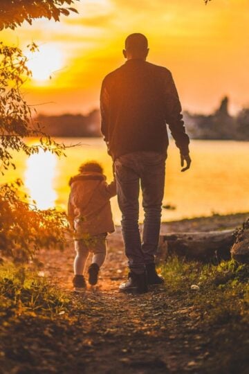 An adult and a child walk hand in hand towards a lake at sunset, surrounded by trees and warm orange light—capturing a perfect moment for those seeking family photography tips.