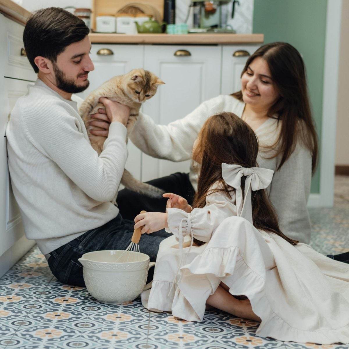 A man holds a cat while a woman and a girl in a white dress sit on a kitchen floor. The girl is whisking something in a large bowl, creating an authentic moment perfect for family photography tips.
