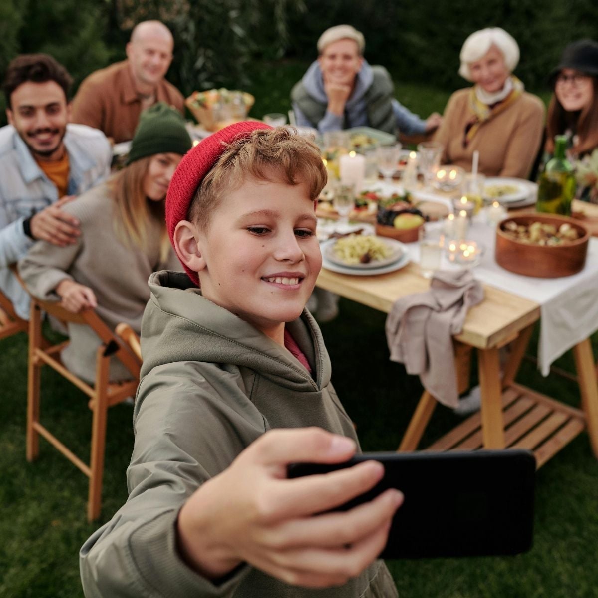 A boy takes a selfie in front of a group of people sitting at an outdoor dining table with food and drinks, capturing a lively moment together and putting family photography tips into practice.