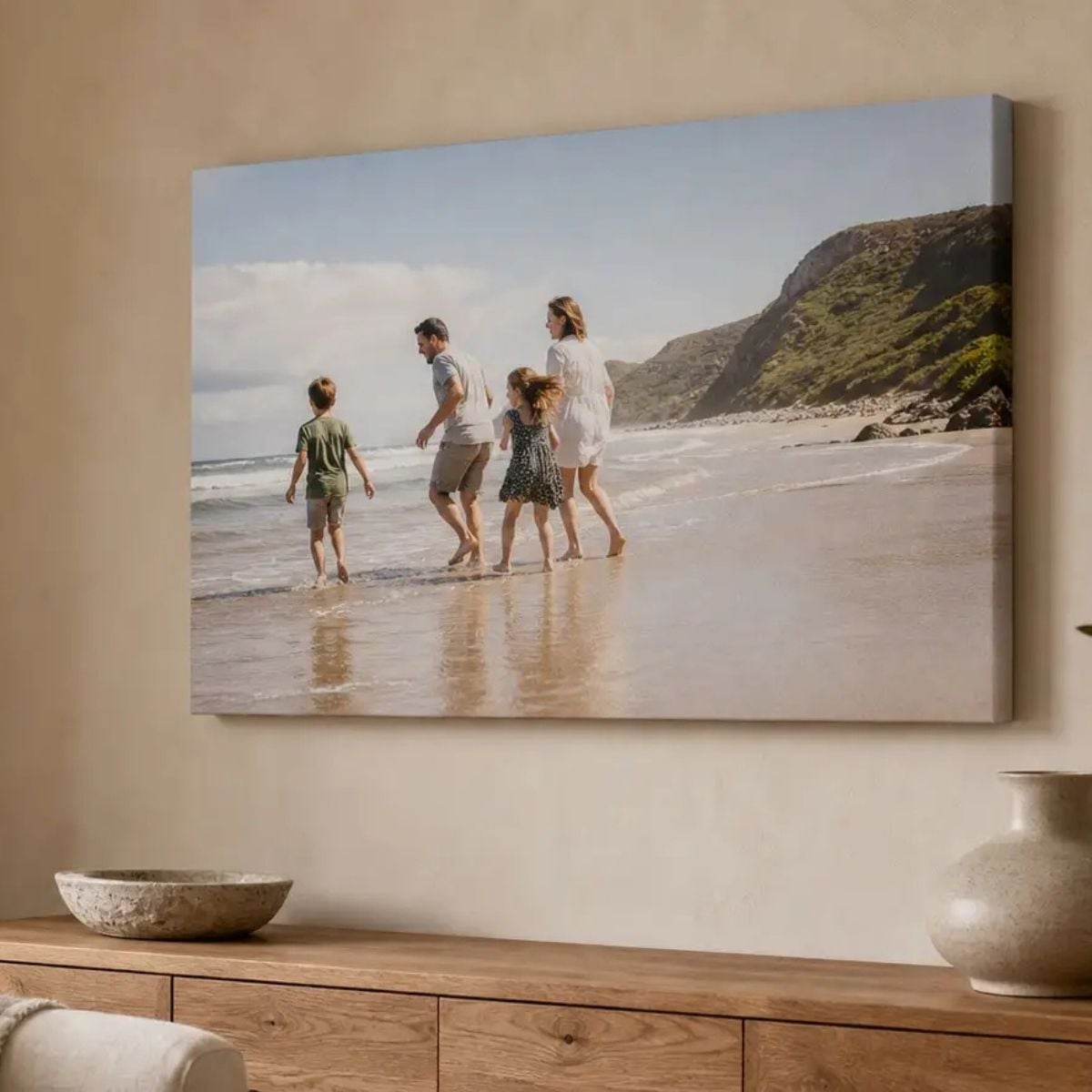 A canvas print on a beige wall shows a family of four walking along a beach near cliffs—a perfect example for anyone seeking family photography tips. A wooden sideboard with a stone bowl and plate sits below the artwork.