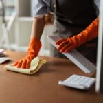 Person wearing orange gloves cleans a computer keyboard with a cloth on a brown desk, highlighting the importance of office cleanliness and maintaining proper office desk hygiene.