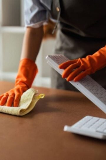 Person wearing orange gloves cleans a computer keyboard with a cloth on a brown desk, highlighting the importance of office cleanliness and maintaining proper office desk hygiene.