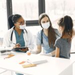 Two female healthcare professionals wearing masks talk to a young girl in a medical surgery, with medicine packs and a clipboard on the table, highlighting the compassionate care provided to children facing paediatric brain cancer and the profound impact on families.