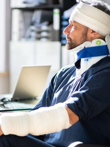 What Makes an Injury Catastrophic? Man with bandages on head and arm, and a neck brace, sitting indoors next to a laptop, looking to the side.