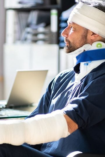 Man with bandages on head and arm, and a neck brace, sitting indoors next to a laptop, looking to the side.