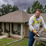 A person wearing safety equipment stands on a ladder clearing leaves from a house gutter, with a brick house and cloudy sky in the background.