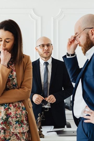 Three people in business attire stand in an office, two looking stressed while one stands in the background observing the scene.