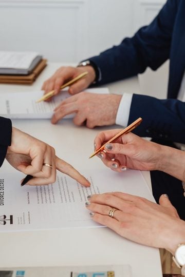 Three people sit at a desk, reviewing and signing a divorce agreement document with legal paperwork and a Lady Justice statuette visible on the table.