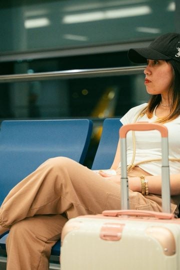 Is It Acceptable to Wear Loungewear to the Airport? We Settle The Debate A woman wearing a black cap and beige loungewear sits on a blue airport bench with her arms folded, next to a pink suitcase.