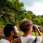 An adult and a child stand outdoors; the child looks through binoculars whilst the adult watches, with trees and greenery in the background, capturing a memorable moment on their luxury safari with an experienced family guide.