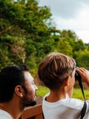 An adult and a child stand outdoors; the child looks through binoculars whilst the adult watches, with trees and greenery in the background, capturing a memorable moment on their luxury safari with an experienced family guide.