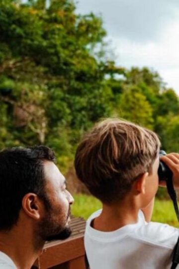 An adult and a child stand outdoors; the child looks through binoculars whilst the adult watches, with trees and greenery in the background, capturing a memorable moment on their luxury safari with an experienced family guide.