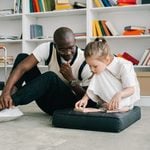 Man and young girl sit on the floor in a library, focused on a wooden puzzle-a setting that fosters support for children as they learn and grow. Bookcases with colourful books are in the background, offering valuable resources and guides for parents navigating behavioural challenges.