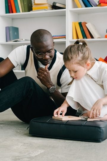 Man and young girl sit on the floor in a library, focused on a wooden puzzle—a setting that fosters support for children as they learn and grow. Bookcases with colourful books are in the background, offering valuable resources and guides for parents navigating behavioural challenges.
