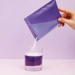A hand pours white powder from a purple packet of Collagen Peptides into a clear glass containing purple liquid, against a light purple background.