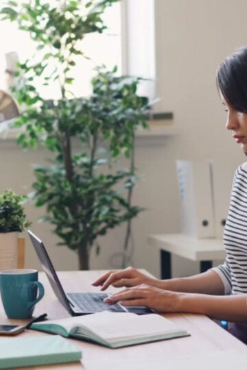 Woman sitting at a desk typing on a laptop, with a mug, notebook, and potted plants nearby, in a bright room with natural light—a peaceful moment that invites mindfulness and encourages her to become more present in her daily routine.