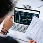 Person holding a financial report and pointing at a laptop screen displaying share charts and market data in an office setting, illustrating the process of starting to trade futures and analyse opportunities in futures trading.