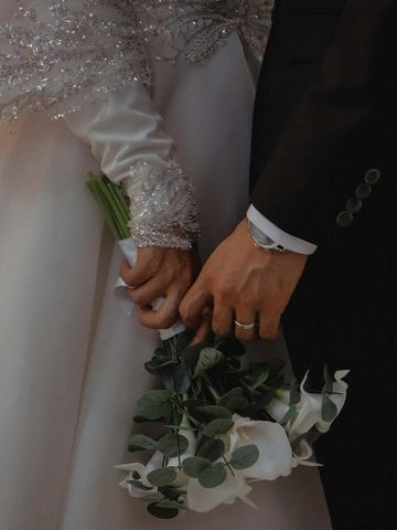 Close-up of a couple holding hands, one in a white, beaded dress holding a bouquet, the other in a black suit wearing a watch and wedding ring.