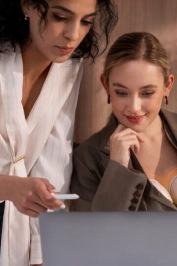 Two women in business attire look at a laptop screen together, one pointing at the screen with a pen, as they discuss the latest advancements in breastfeeding technology, including a smart breast pump with app connectivity, in an office setting.